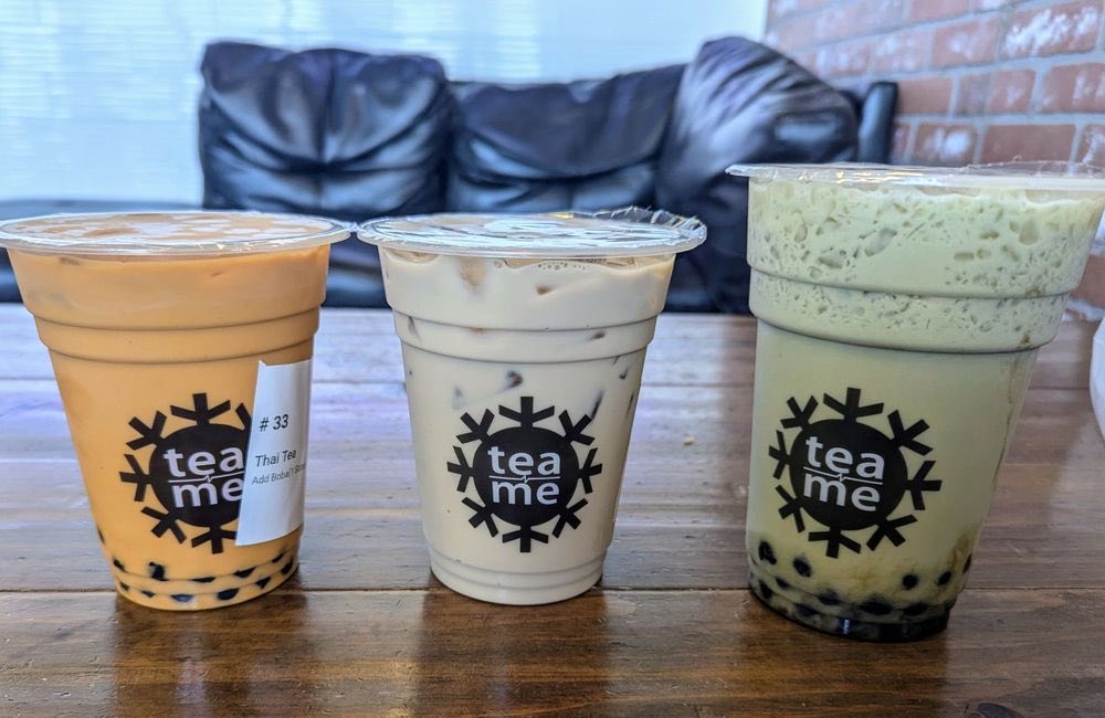 Three iced boba drinks in Tea N' Me cups lined up on a wooden table, with a black leather couch and a brick wall behind them.