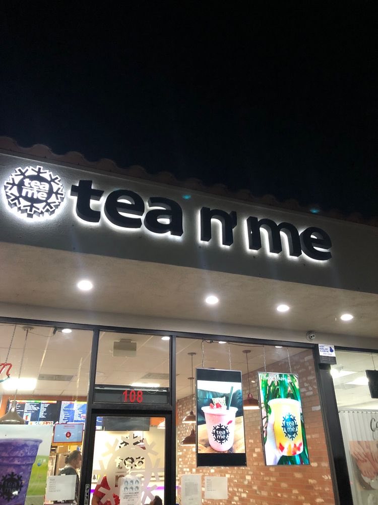 The lit Tea N' Me sign above the shopfront at night, with the windows glowing inside.