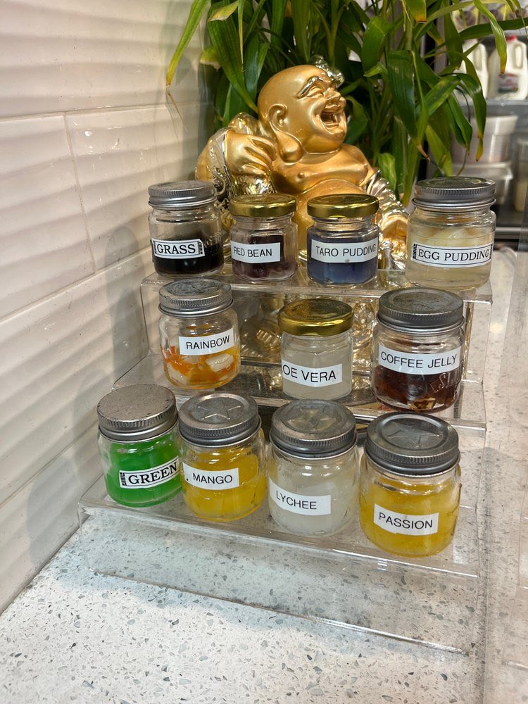 Glass jars of toppings on a tray by the counter, labelled grass jelly, red bean, taro pudding, egg pudding, rainbow, aloe vera, coffee jelly, mango, lychee and passion.